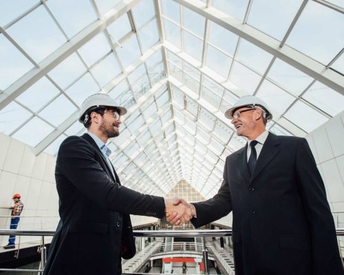 two-cheerful-businessman-handshaking-helmet-new-building (1)